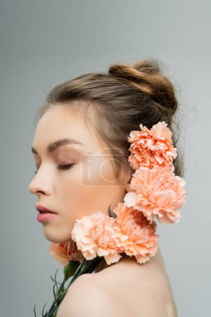 young woman with closed eyes and natural makeup near fresh carnations isolated on grey