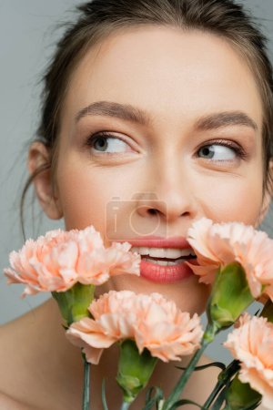portrait of smiling woman with natural visage looking away near fresh carnations isolated on grey