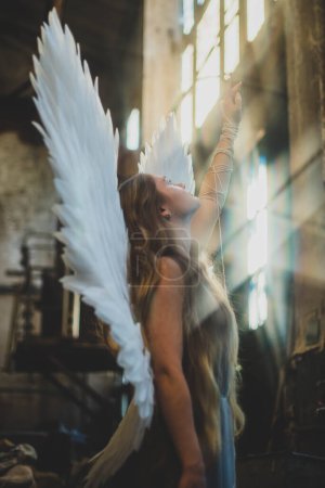 天使女孩站在凌乱的车间里.一个背带翅膀的女孩向太阳伸出手来