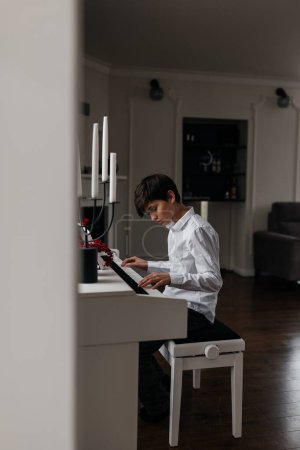 英俊的12岁男孩穿着白衬衫弹奏数字钢琴。在家里练习。家庭音乐会