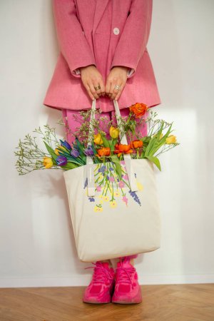 穿着粉色衣服的时髦女人把装满鲜花的手提包放在室内，用手提包进行特写。春天、风格和生态购物袋概念