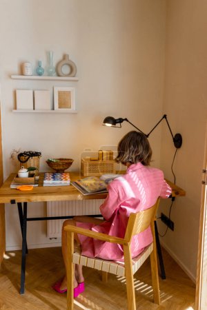 穿着粉色西服的女人坐在时尚时尚的现代公寓工作室的工作区边看书