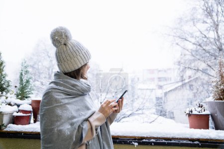 一个戴着温暖帽子和毛毯的女人手里拿着智能手机。背景为降雪的冬季景观.
